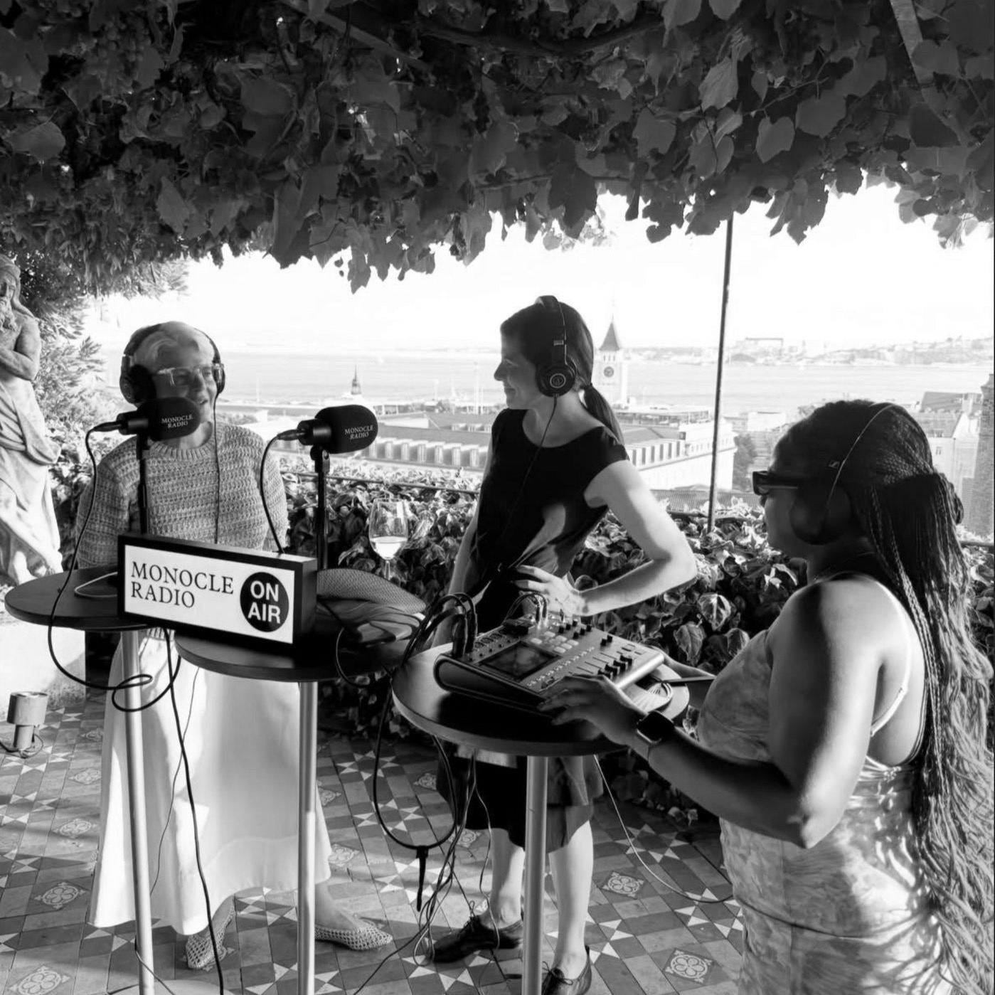 Monocle Radio on air under a vine-covered pergola above the Tagus in Lisbon: a presenter and guest at the microphones, a producer at the mixing desk